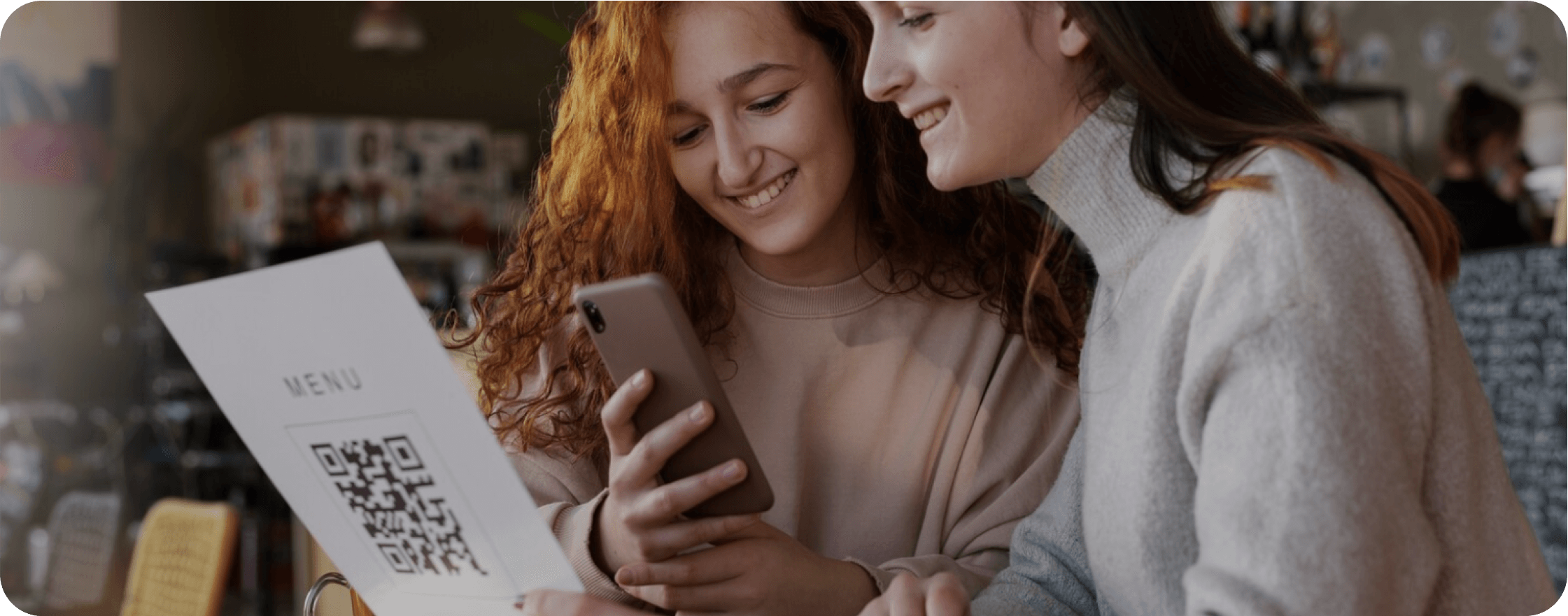 Two girls are sitting at the restaurant, one of them looks at the menu, the other one on her smartphone.