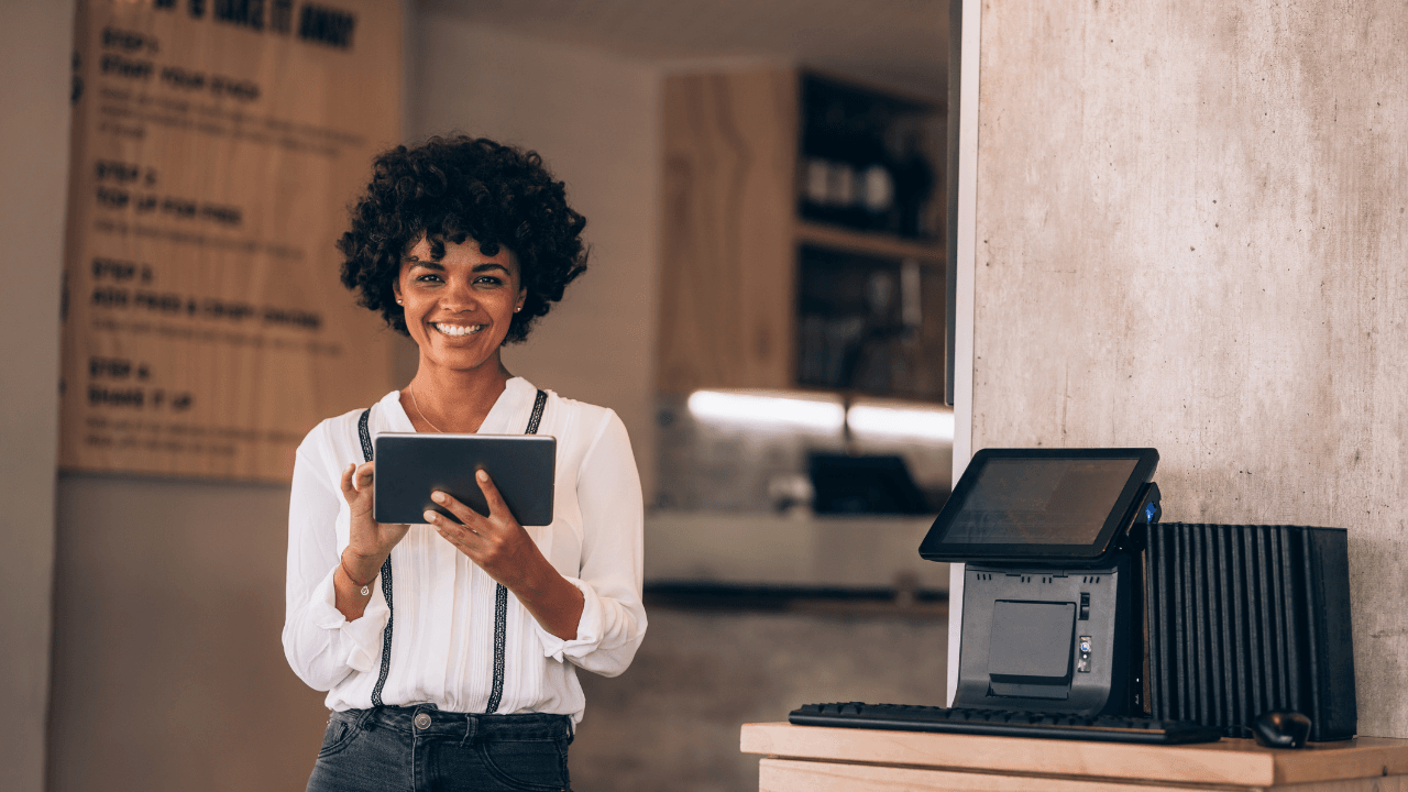 A curly-haired young woman holds a tablet and smiles at the camera.