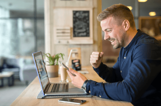 A happy man is sitting with his laptop and looking at the smartphone.