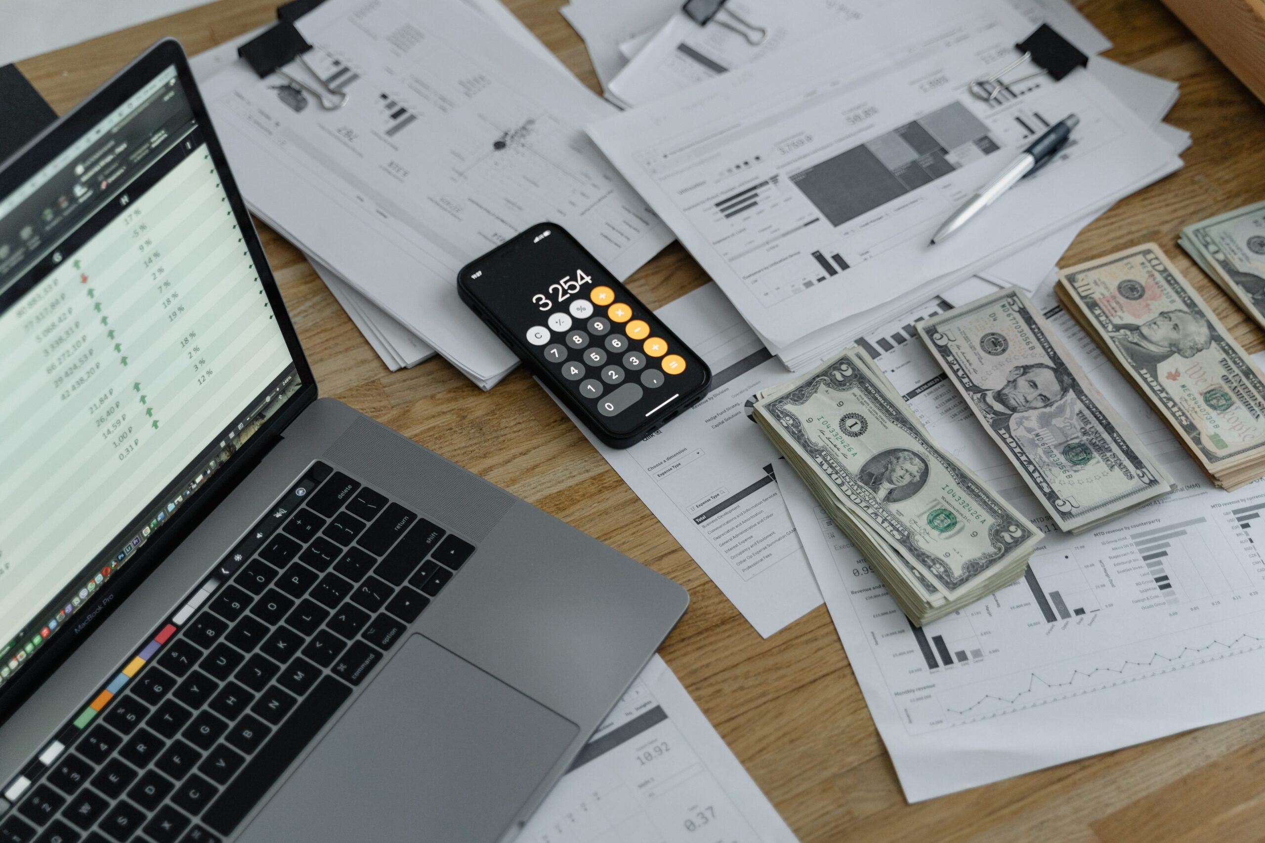 Money, papers and iPhone is placed on the table next to a laptop.